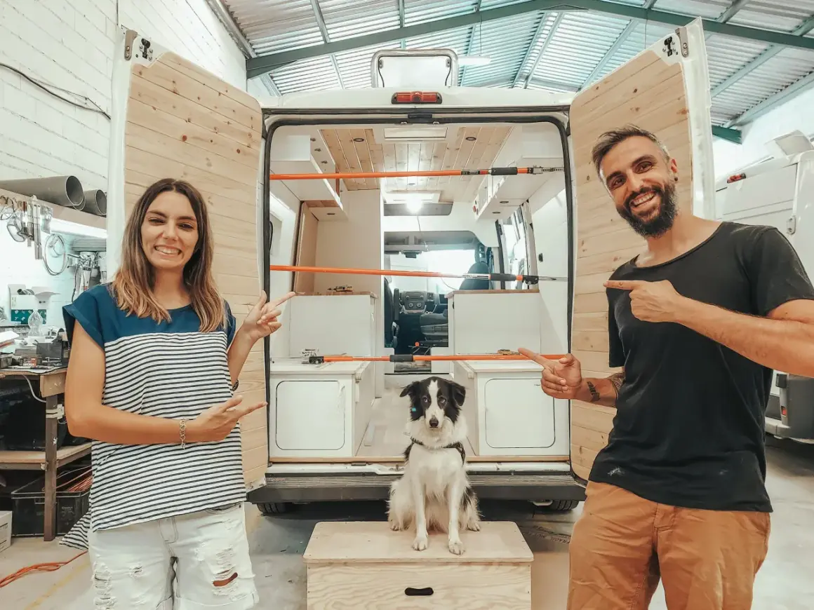 Pareja sonriente y su perro posan junto a una furgoneta camper en proceso de construcción, ¡las mejores furgonetas camper calidad precio!