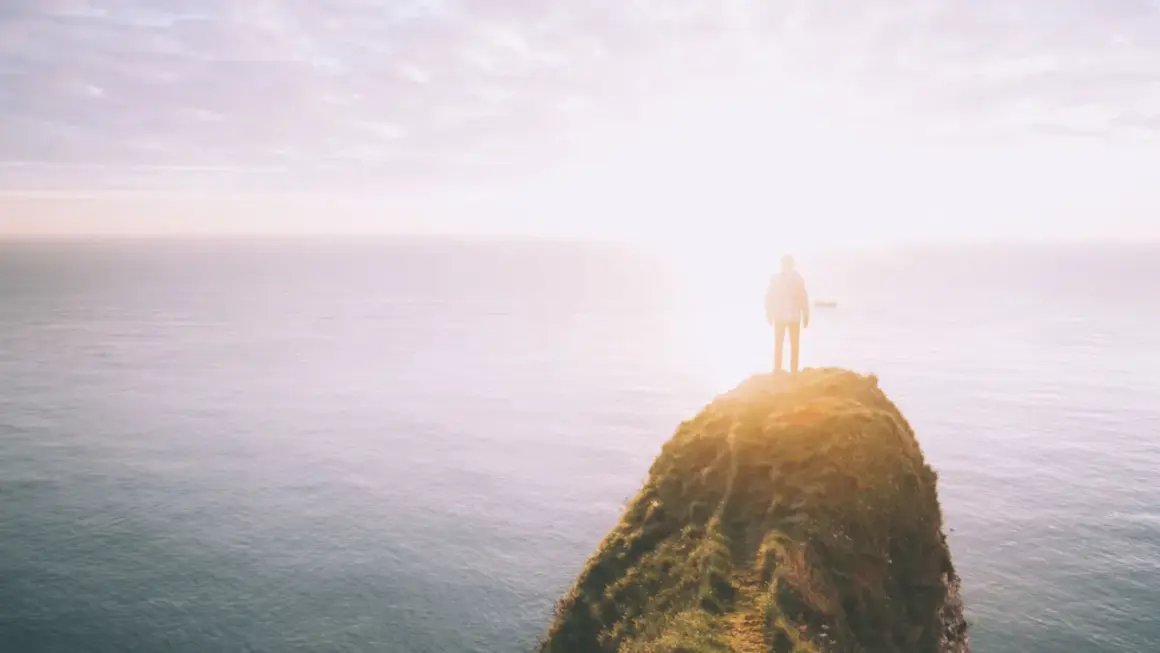 Ein Mensch steht auf einer Klippe am Meer, die Sonne scheint hell. Ein Symbol für das Durchhalten in schweren Zeiten, wenn man dem Horizont entgegenblickt.