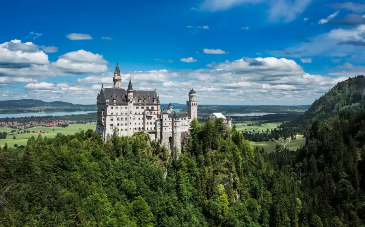 El castillo del rey loco, Neuschwanstein, se alza majestuoso sobre un risco verde, con torres que acarician las nubes y un paisaje de ensueño.