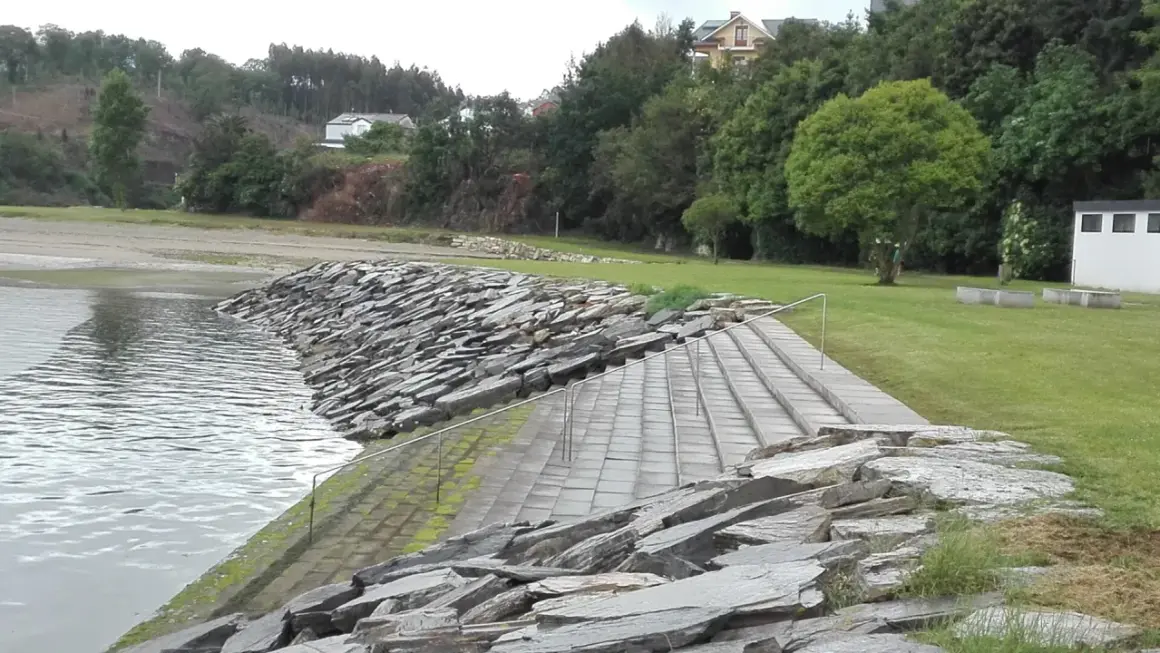 Escaleras de piedra y césped verde junto al agua en la playa de los bloques Ribadeo, con casas y árboles al fondo.