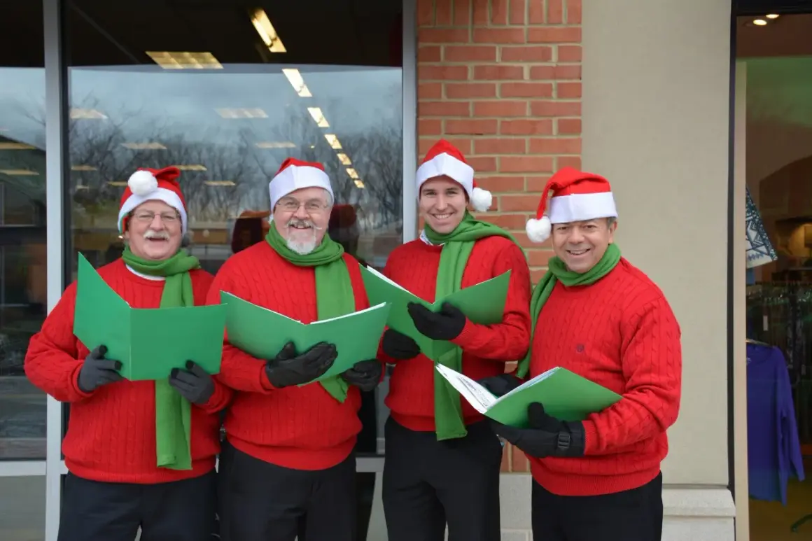 Cuatro hombres con gorros de Papá Noel y bufandas verdes cantan villancicos en inglés famosos, sosteniendo partituras verdes.