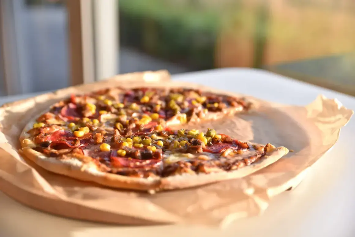 Pyszna pizza z kukurydzą, salami i oregano, podana na papierze do pieczenia, w słonecznym świetle.