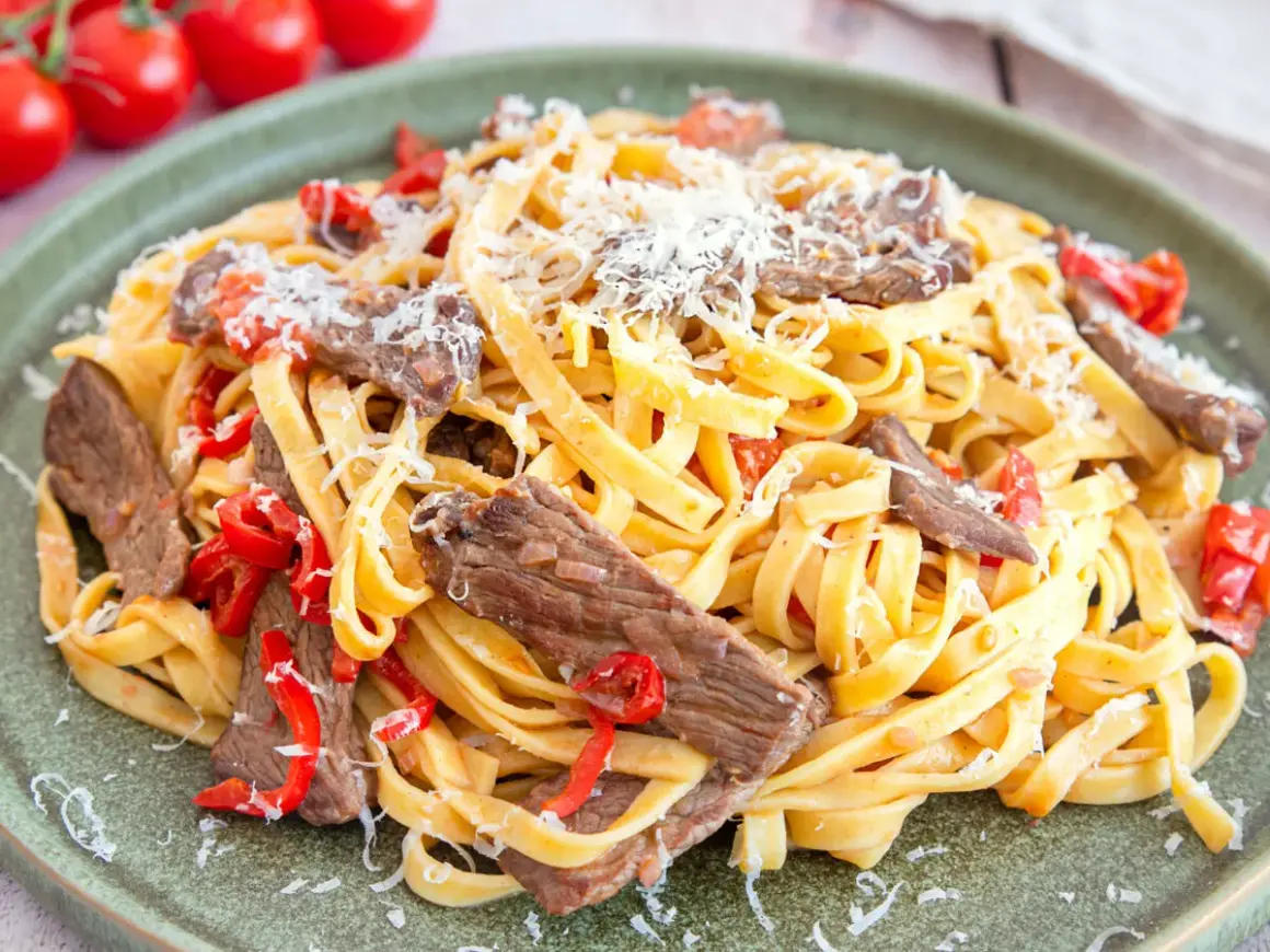 Herzhaftes Gericht mit Pasta und zarten Rinderfiletstreifen, garniert mit geriebenem Parmesan und roten Paprikastücken.