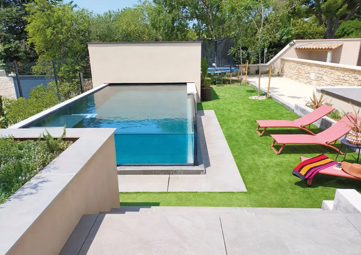 Un magnifique bassin vitré aux eaux bleues, entouré d'une pelouse synthétique et de chaises longues roses.