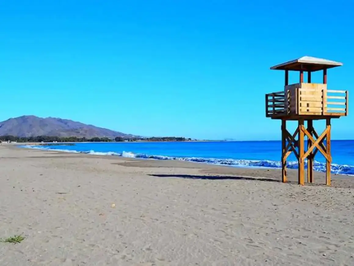 Torre de vigilancia en la vera playa, con el mar azul y montañas al fondo.