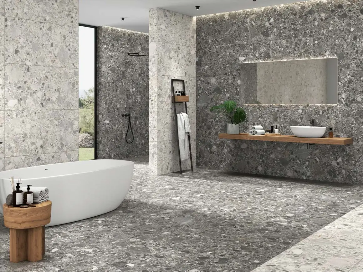Baño moderno con bañera exenta y lavabo sobre encimera. Las paredes y el suelo muestran diferentes tipos de azulejos con efecto terrazo en tonos grises.