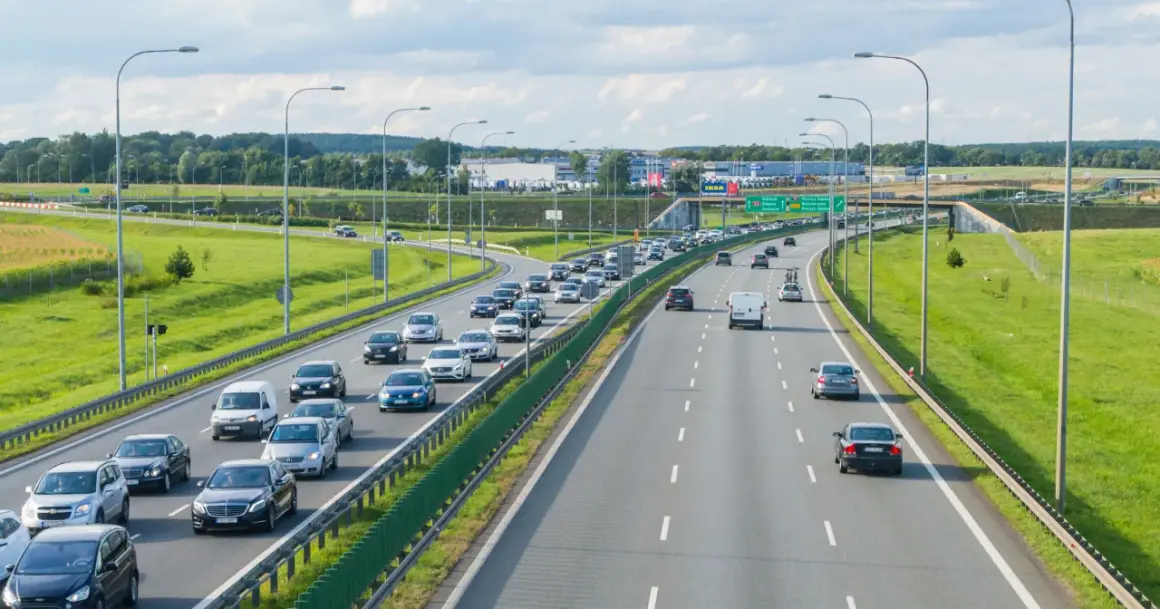 Autostrada z ruchem samochodowym w słoneczny dzień, z zielonymi poboczami i drzewami w tle.