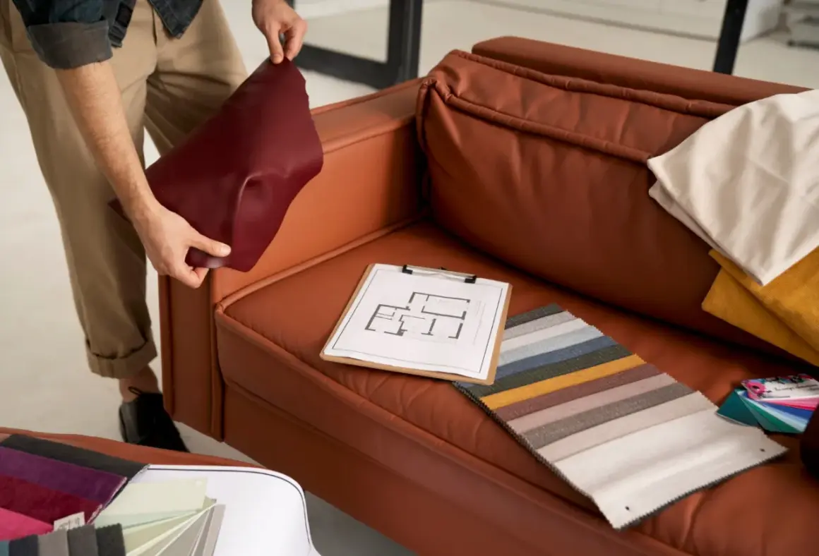 A person is choosing couch materials, holding a burgundy swatch next to a brown leather couch. Fabric samples and a floor plan are on the seat.