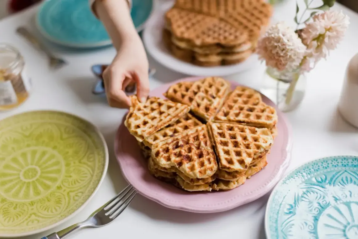 Ein Kind greift nach einem herzförmigen Waffel ohne Ei. Mehrere Stapel Waffeln liegen auf rosa und weißen Tellern, umgeben von bunten Tellern und Blumen.