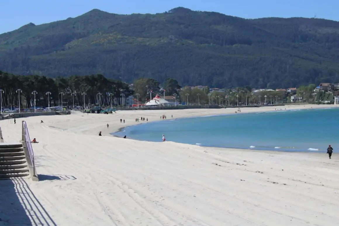Amplia playa de arena blanca en la playa de Samil, con aguas turquesas y una montaña verde al fondo.