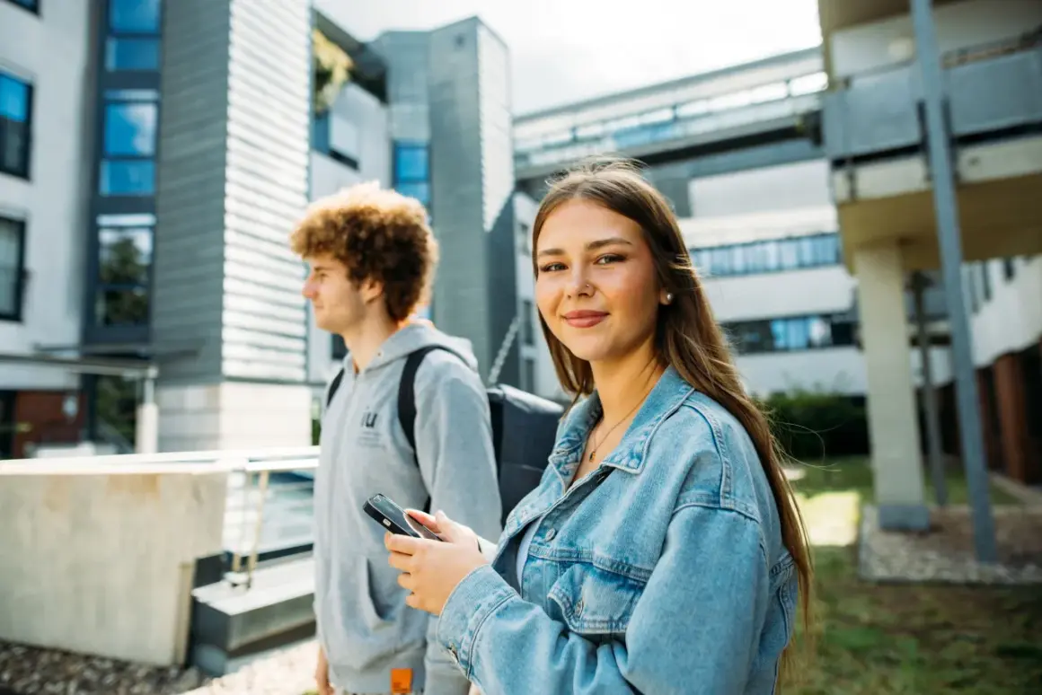 Zwei Studierende vor moderner Architektur. Sie lächelt in die Kamera, er blickt zur Seite. Ist die IU privat?