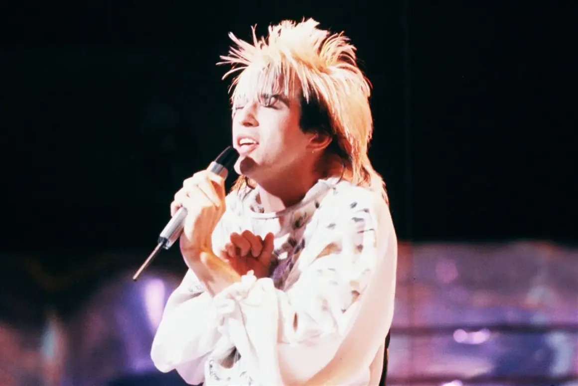 Limahl piosenkarz z charakterystyczną fryzurą śpiewa do mikrofonu na scenie.