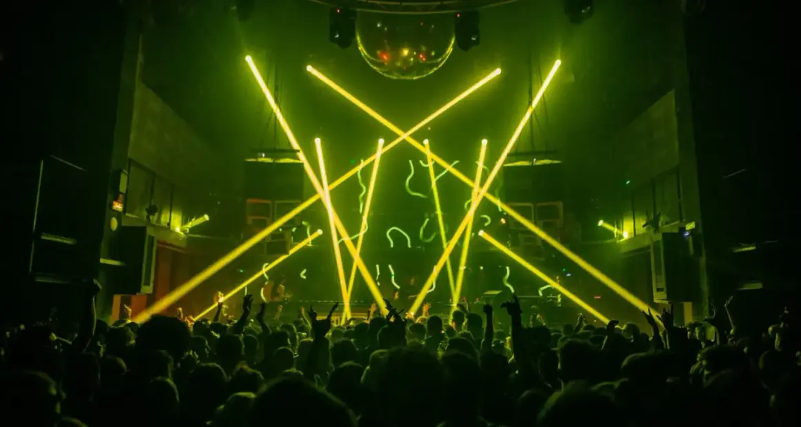 Folla in delirio in un techno club, illuminato da fasci di luce gialla e una sfera stroboscopica.