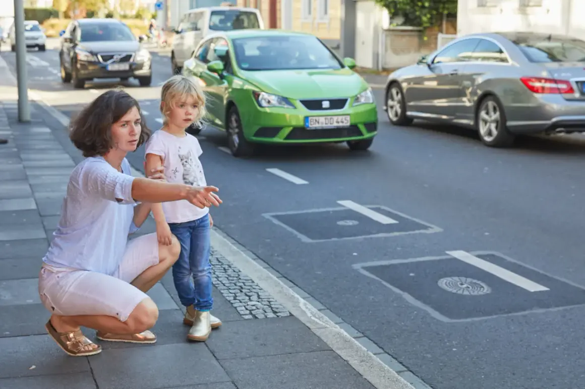 Mutter erklärt ihrer Tochter, dass sie auf dem Gehweg nicht in falsche Richtung fahren darf. Ein grünes Auto fährt auf der Straße.