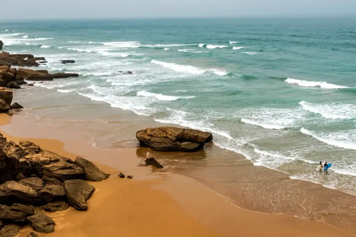 Maroko morze. Dwie osoby z deskami surfingowymi wchodzą do wody na piaszczystej plaży, fale rozbijają się o skaliste wybrzeże.