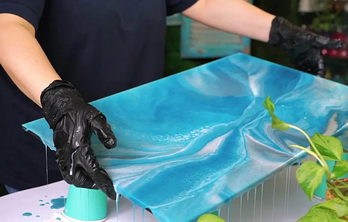 Mani guantate controllano un'opera d'arte in resina epossidica blu e bianca, simile al mare. Il tempo di indurimento della resina epossidica è cruciale per questo effetto.