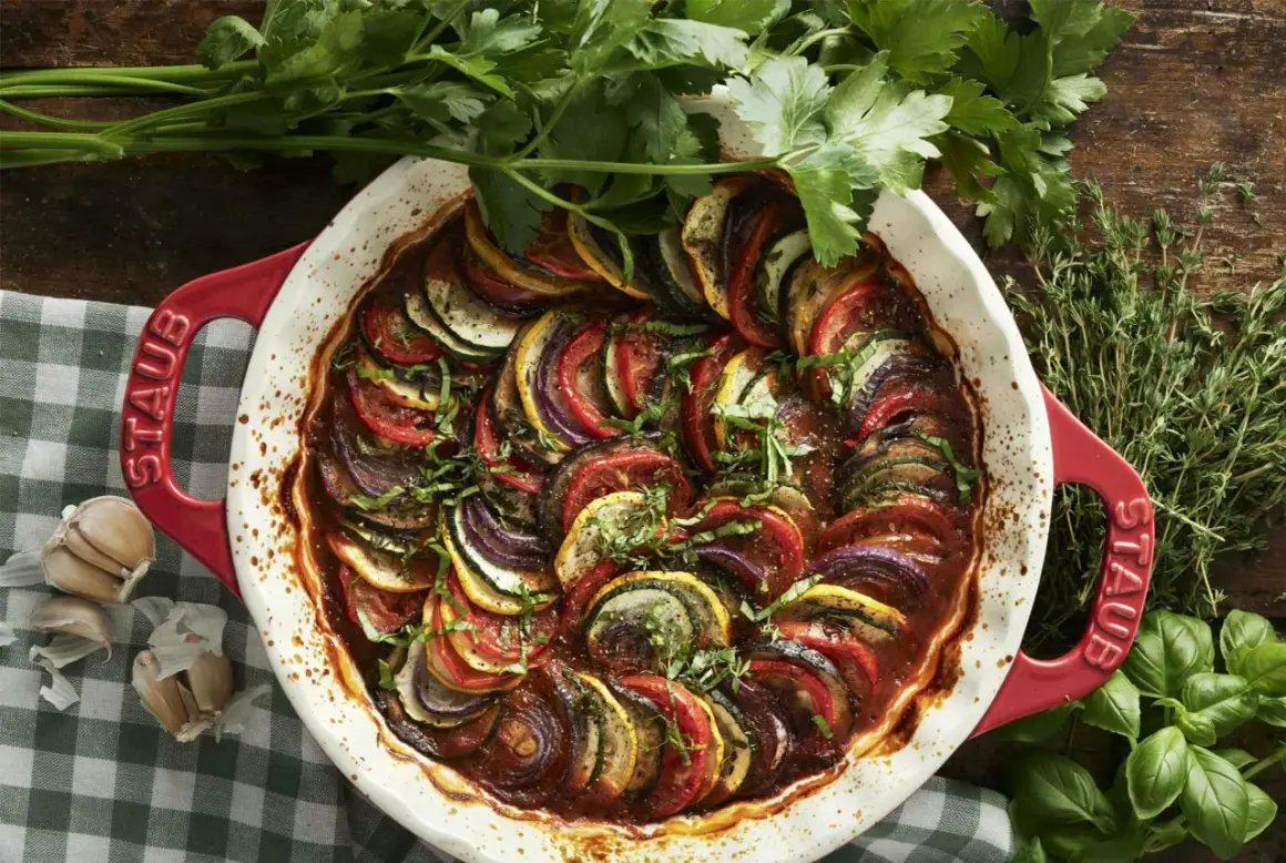 Vacker ratatouille i en röd ugnsform, med skivade grönsaker som tomat, zucchini och aubergine. Perfekt för ett ratatouille recept.