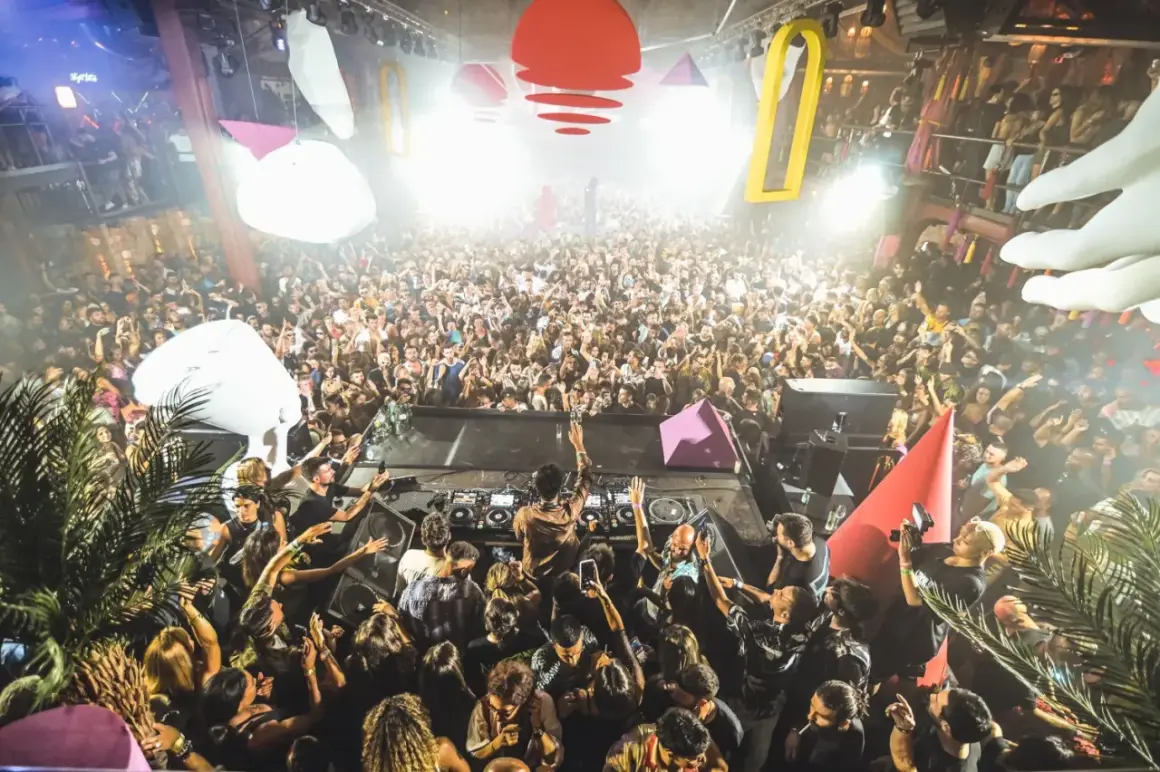 Una folla immensa balla al ritmo di una **canzone ibiza** in un locale notturno. Luci stroboscopiche e decorazioni colorate creano un'atmosfera elettrizzante.