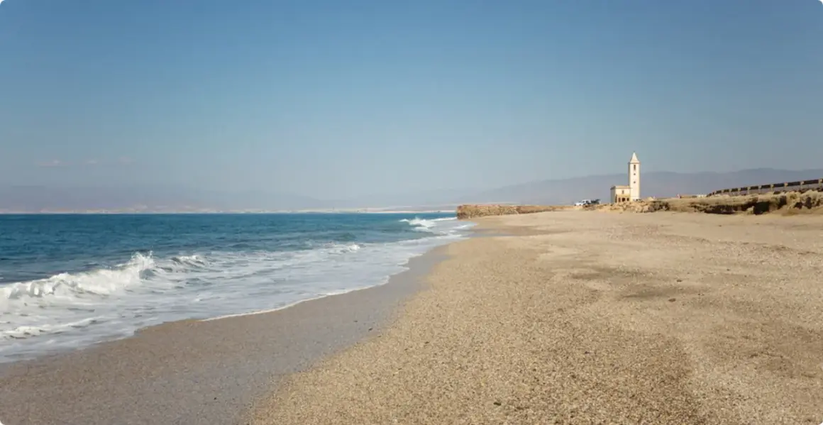 La almadraba de Monteleva, una torre blanca se alza en la playa, con el mar azul y las montañas al fondo.