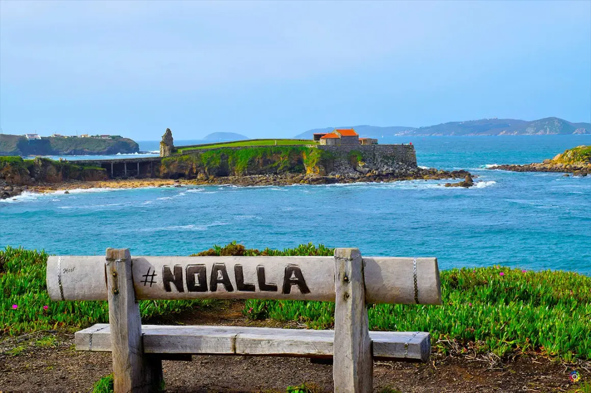 Banco de madera con #NOALLA grabado, frente al mar azul y un pueblo costero con edificios históricos.