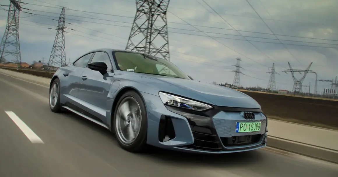 Szary, sportowy Audi GT jedzie po drodze, a w tle widać linie energetyczne i słupy.