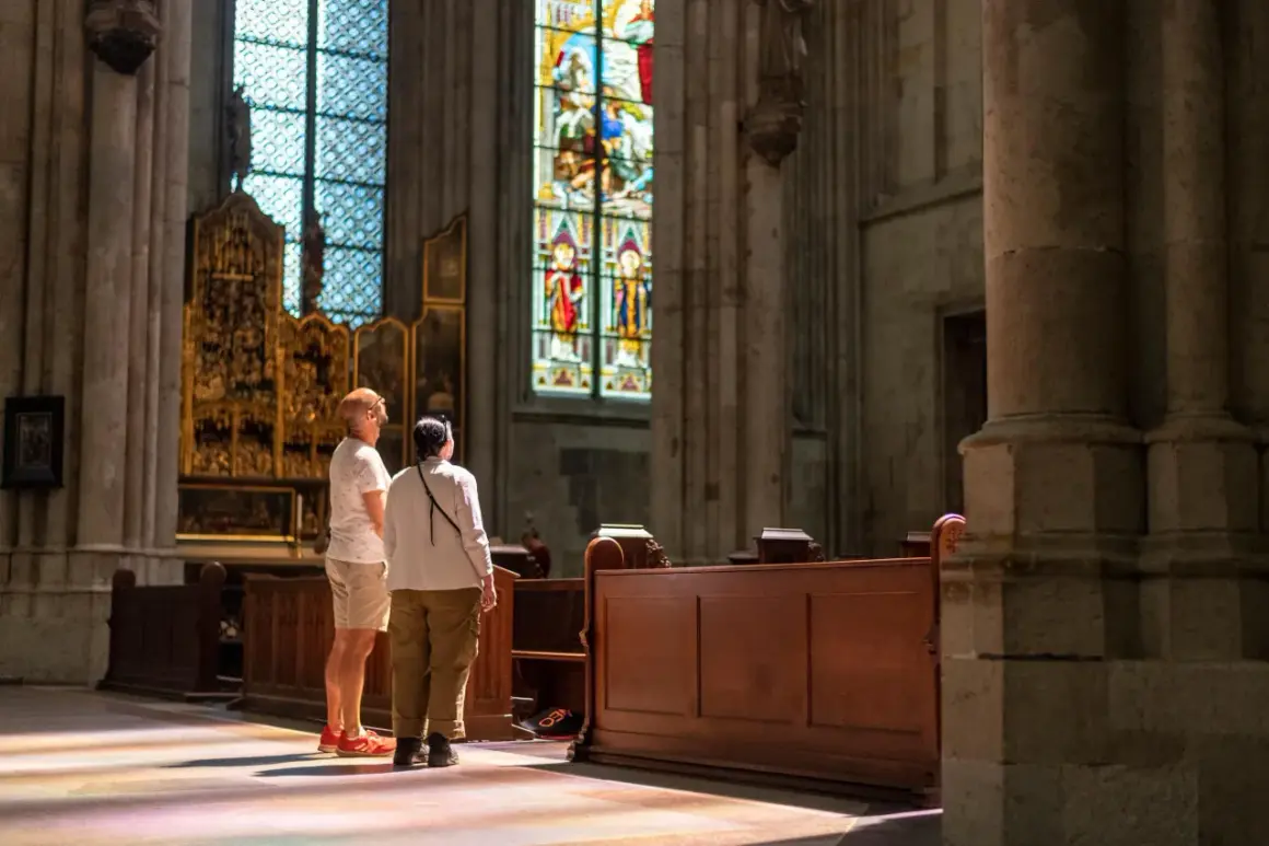 Zwei Besucher bestaunen die bunten Glasfenster im Kölner Dom. Sonnenlicht fällt durch die Fenster und malt bunte Muster auf den Boden.