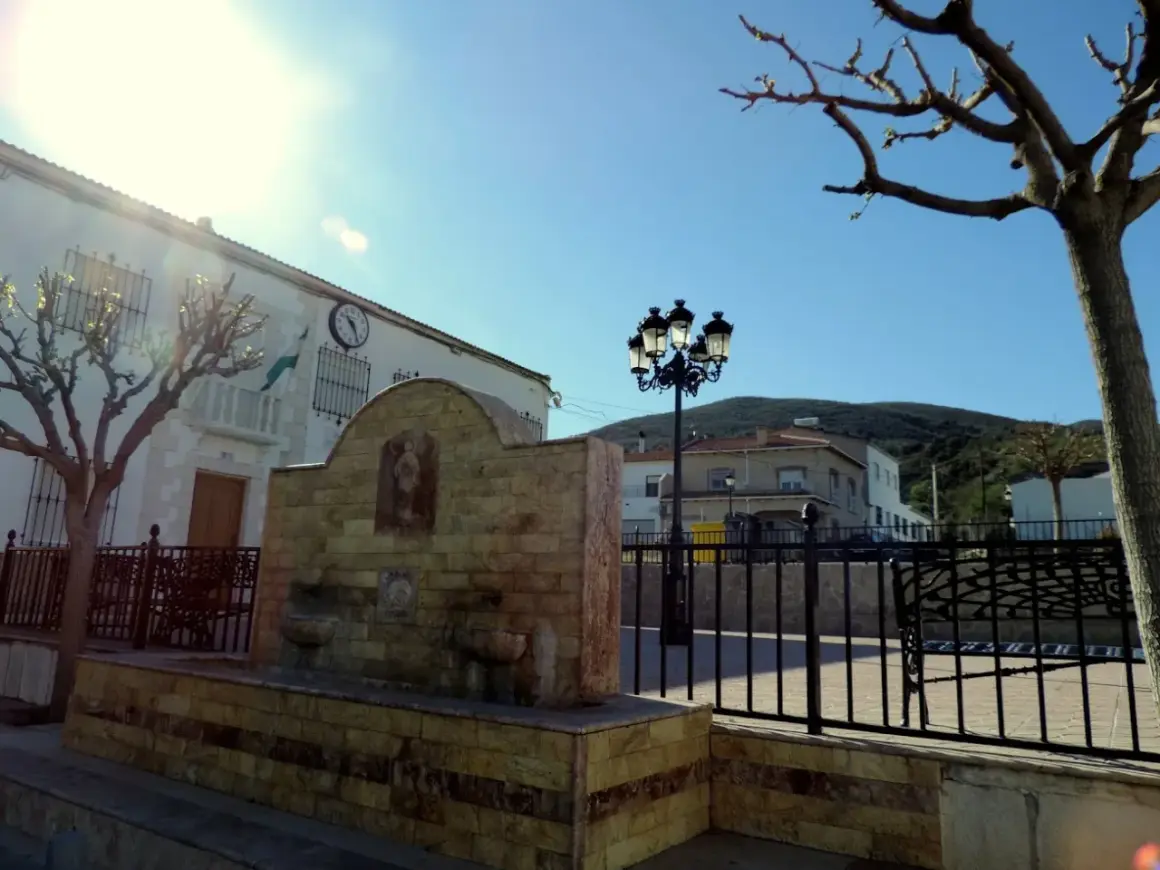 Fuente de piedra en Chercos, Almería, con un edificio blanco y un reloj al fondo.