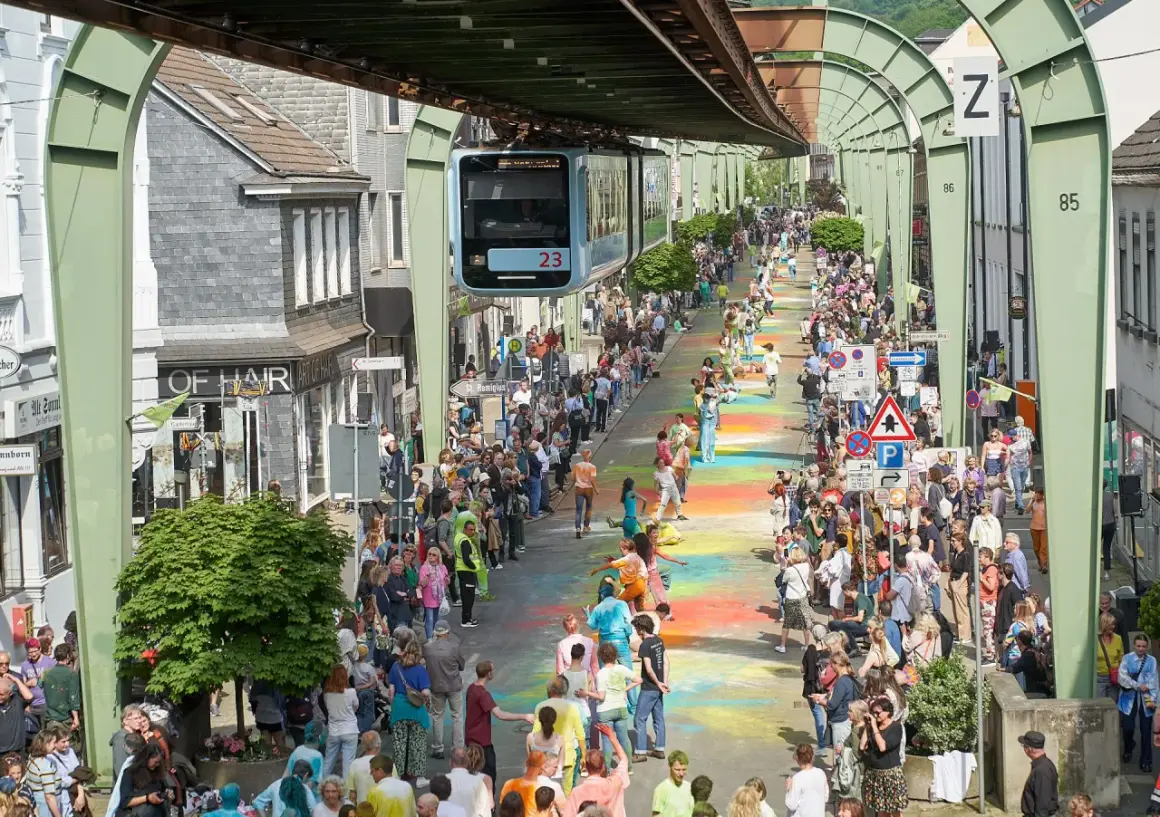 Bunte Farben auf dem Boden und eine Schwebebahn über der belebten Straße im Wuppertal Zentrum. Menschenmassen säumen die bunte Gasse.