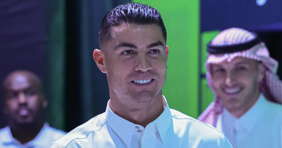 Cristiano Ronaldo w białej koszuli, uśmiecha się. W tle mężczyzna w tradycyjnym arabskim stroju.