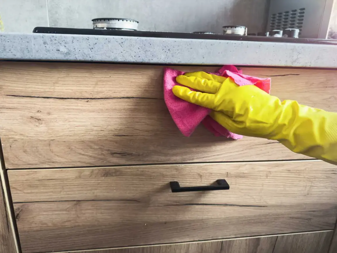 Main jaune essuyant une tache sur bois avec un chiffon rose.