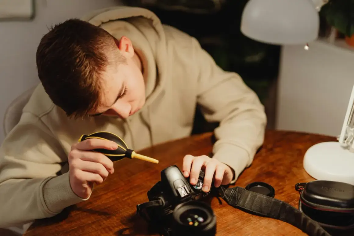 A young person carefully performs camera cleaning, using a blower to remove dust from their DSLR.