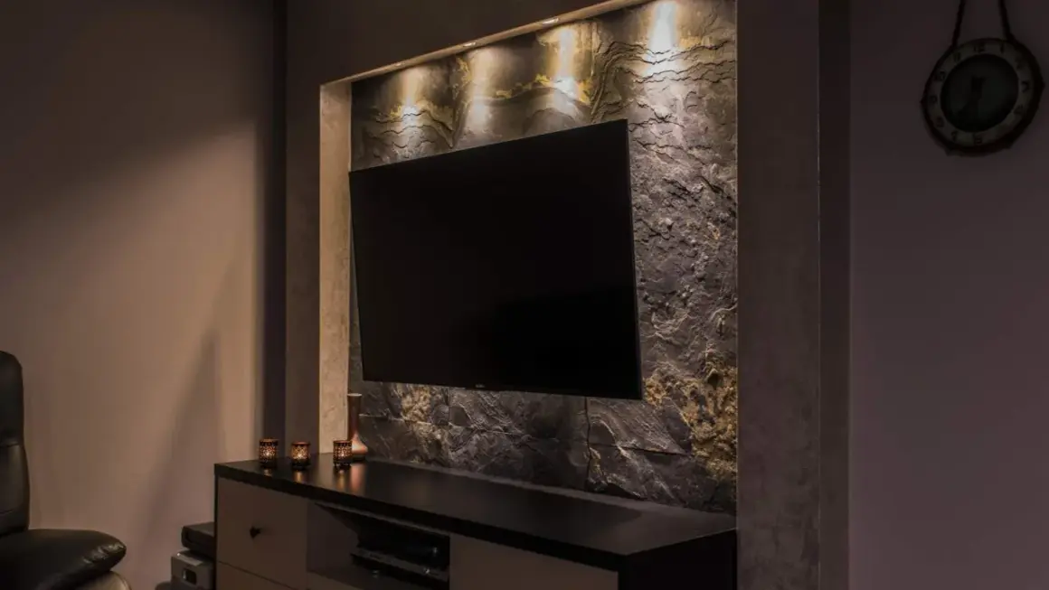 Nowoczesny salon z telewizorem wiszącym na ścianie wyłożonej kamieniem łupek. Oświetlenie podkreśla fakturę kamienia.