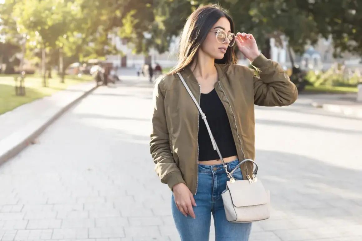 Młoda kobieta w stylowej kurtce wiosennej khaki, jeansach i z torebką na ramieniu spaceruje po parku.