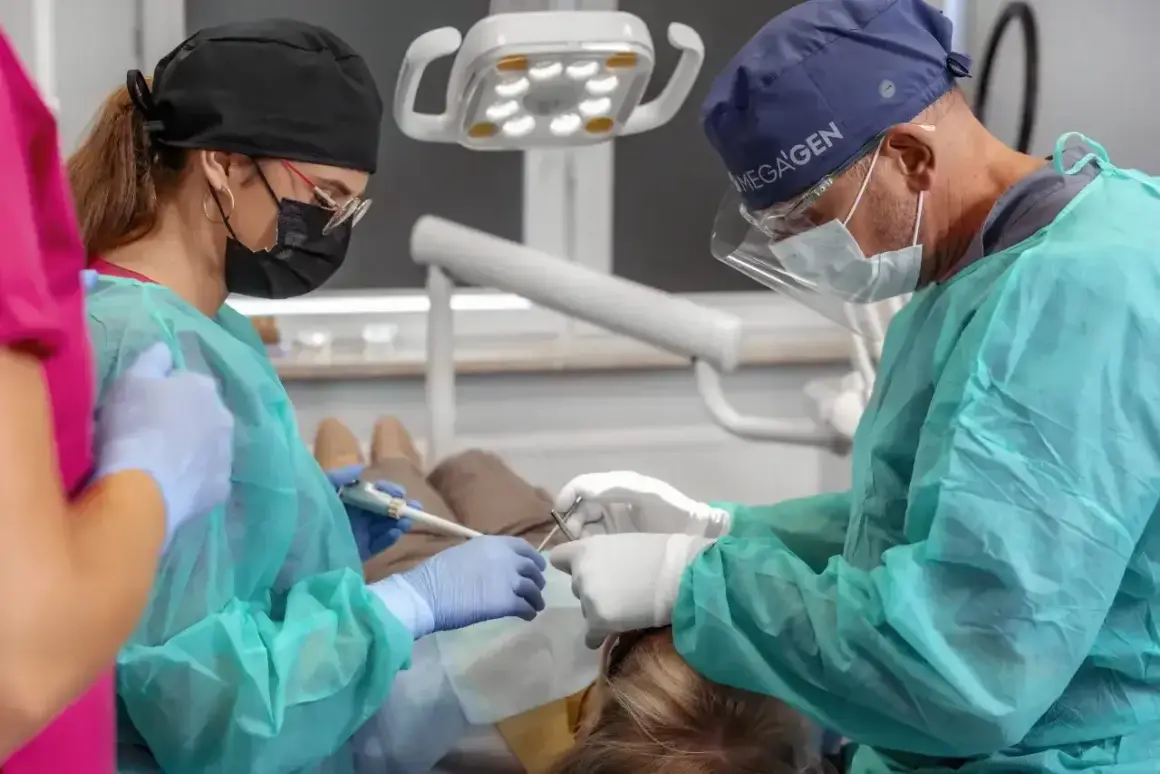 Dentysta i asystentka w maseczkach i fartuchach wykonują zabieg. Widać narzędzia i lampę zabiegową.