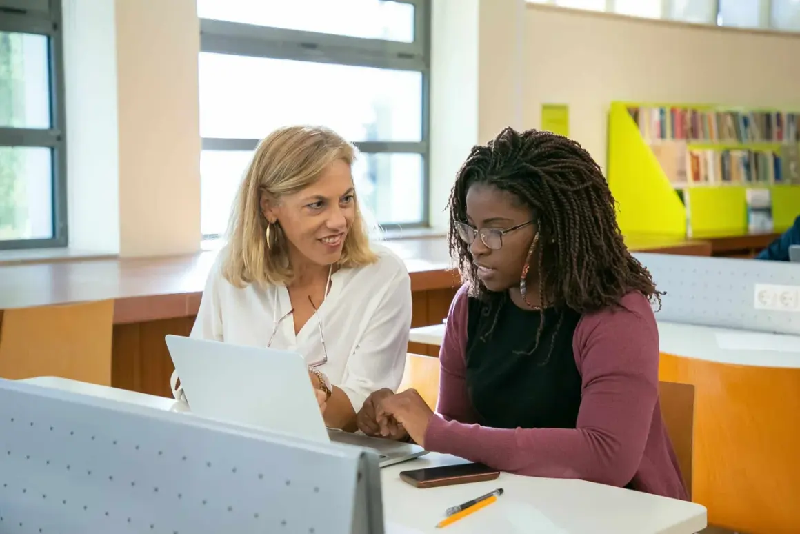 Dos mujeres en un aula, una rubia y otra afrodescendiente, revisan un portátil. La rubia sonríe, la otra parece preocupada, quizás porque su aptis caduca.