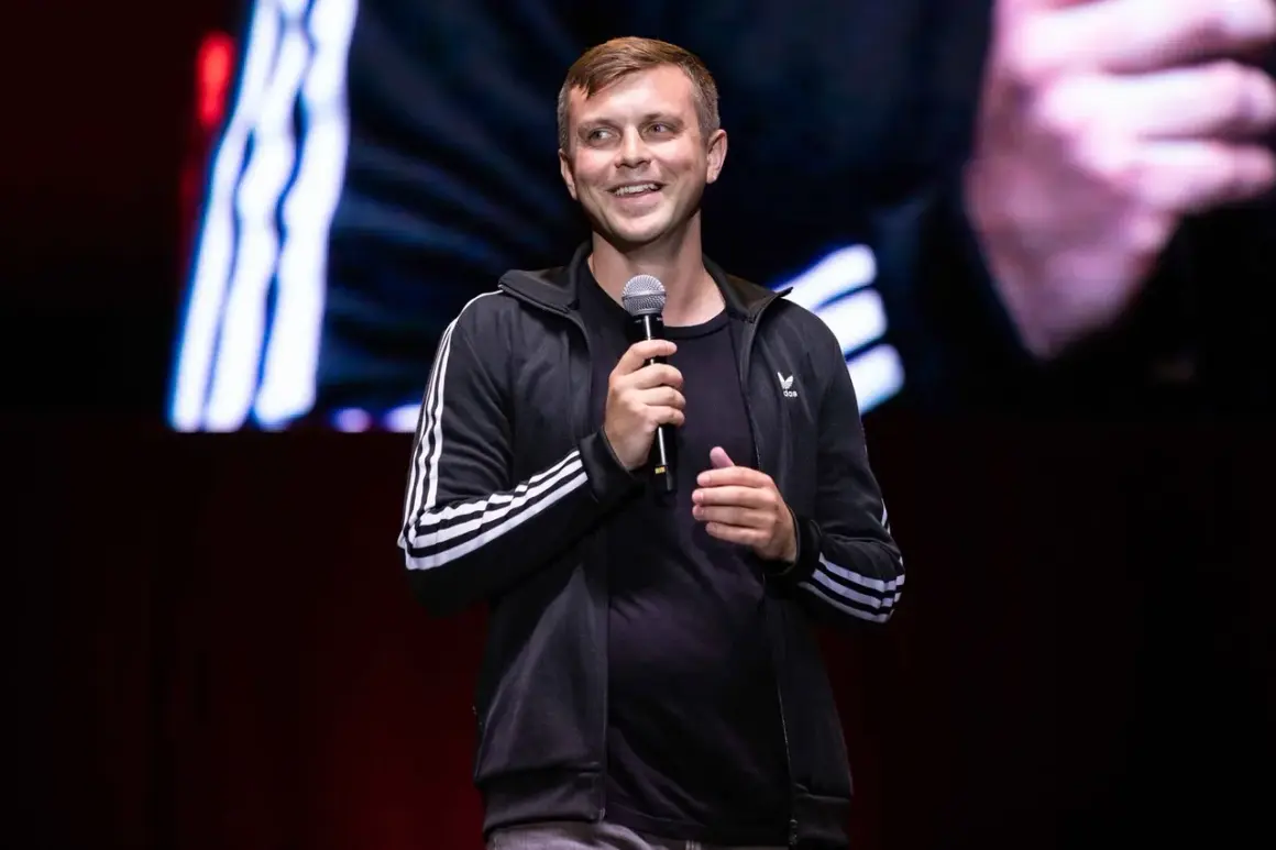 Uśmiechnięty Krajewski na scenie, trzyma mikrofon. Jego stand up rozgrzewa publiczność.