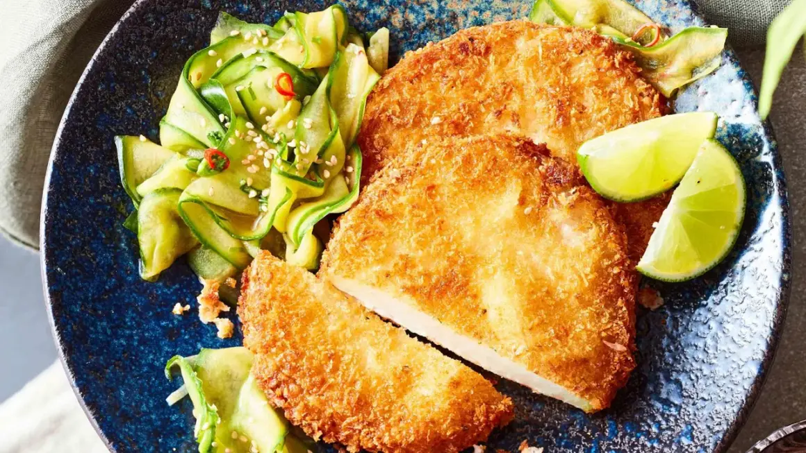 Knuspriges Kohlrabi-Schnitzel mit Gurkensalat und Limettenspalten auf einem blauen Teller.