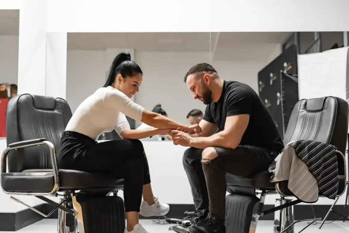 Barber w czarnym t-shircie i podartych jeansach siedzi na fotelu, trzymając dłoń klientki. Zastanawia się, ile zarabia barber.