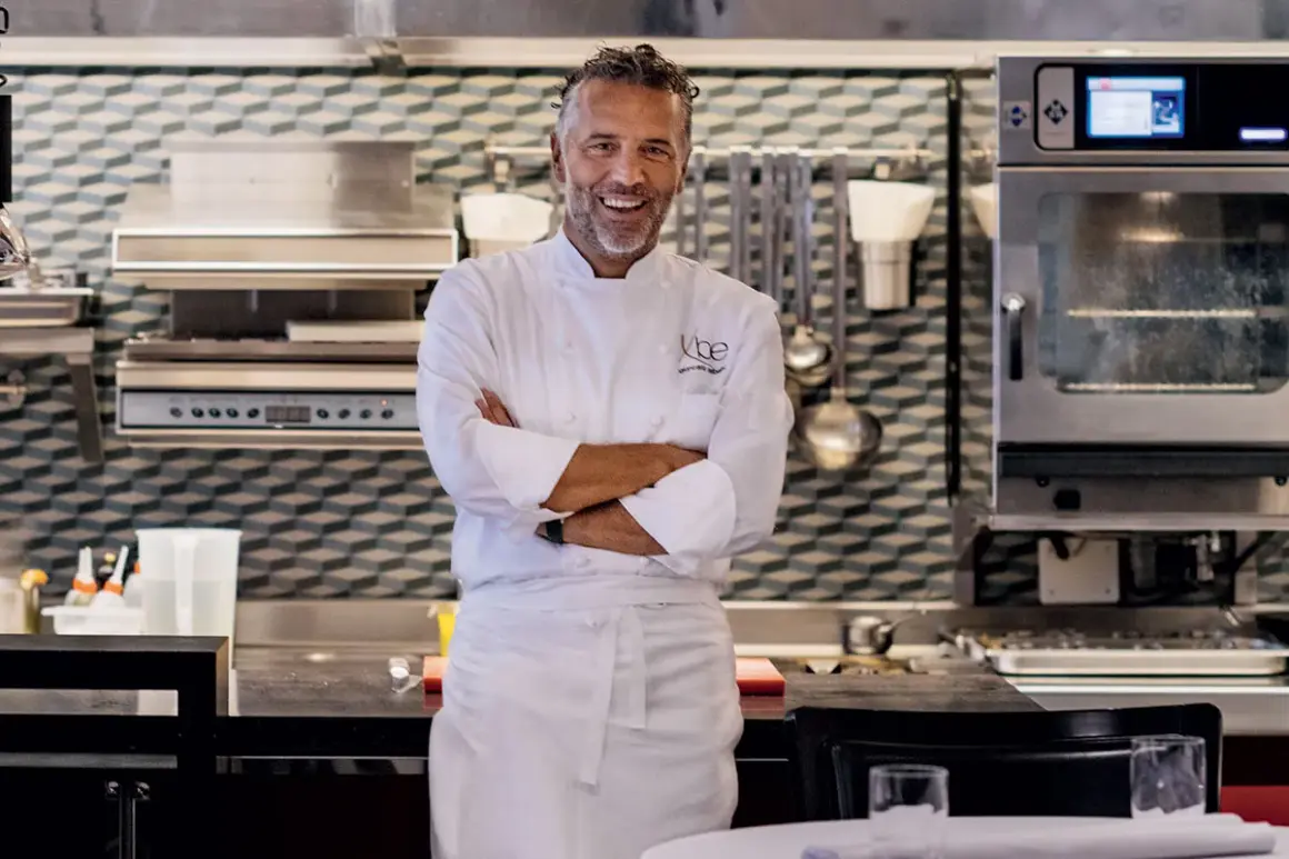 Chef Perbellini, sorridente in cucina, con le braccia conserte. Dietro di lui, attrezzature professionali e un forno acceso.