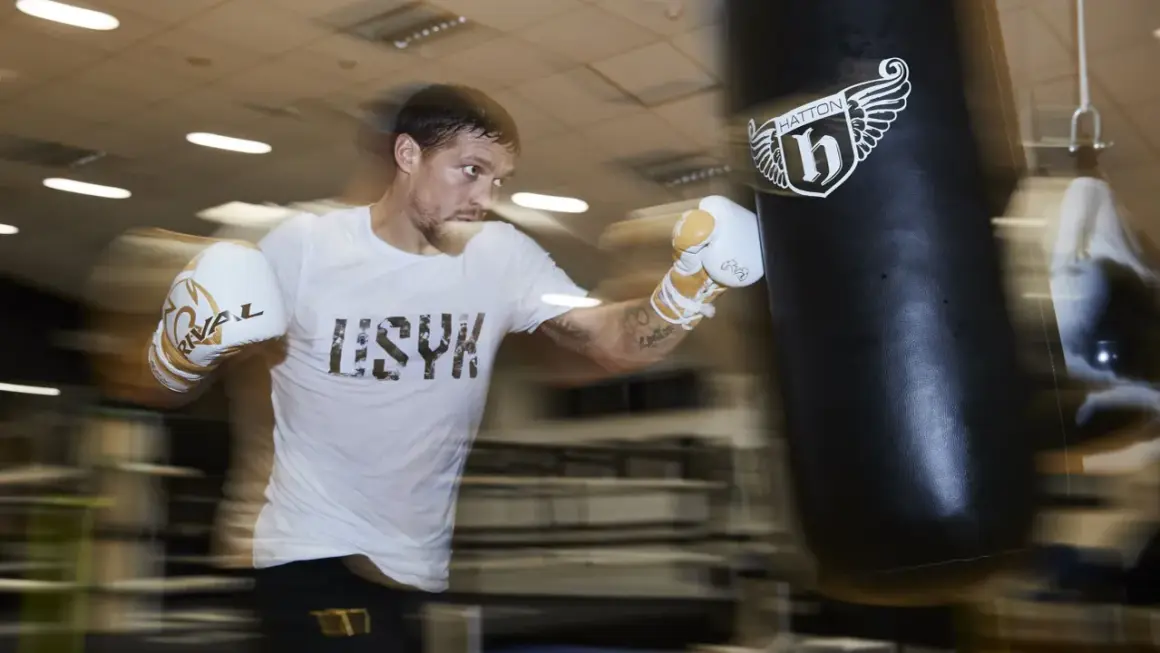 A boxer in a white t-shirt and gloves trains intensely during a boxing training camp, punching a heavy bag.