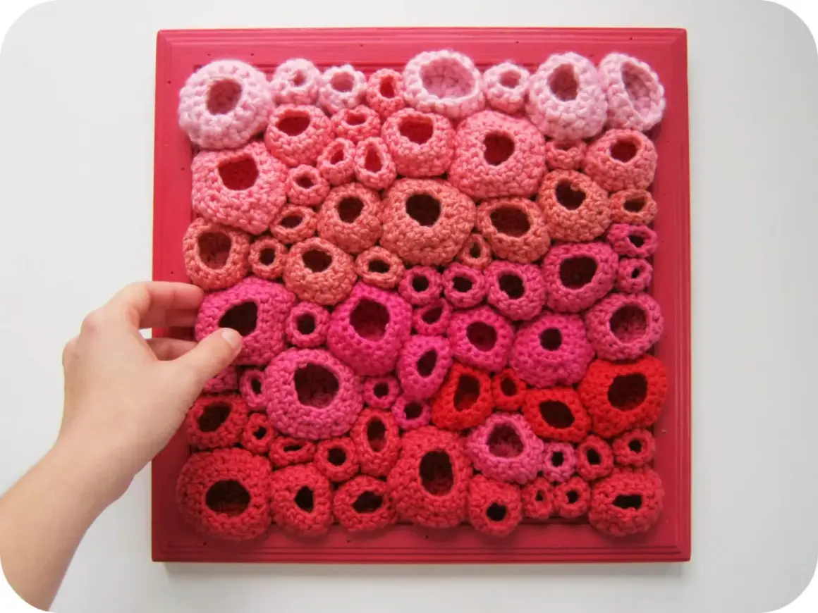 Opera d'arte in freeform crochet con forme tondeggianti rosa e rosse su telaio rosso. Una mano sfiora le creazioni.