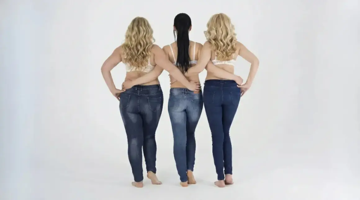 Tre donne in jeans, una con fisico a pera uomo, mostrano diverse forme del corpo.
