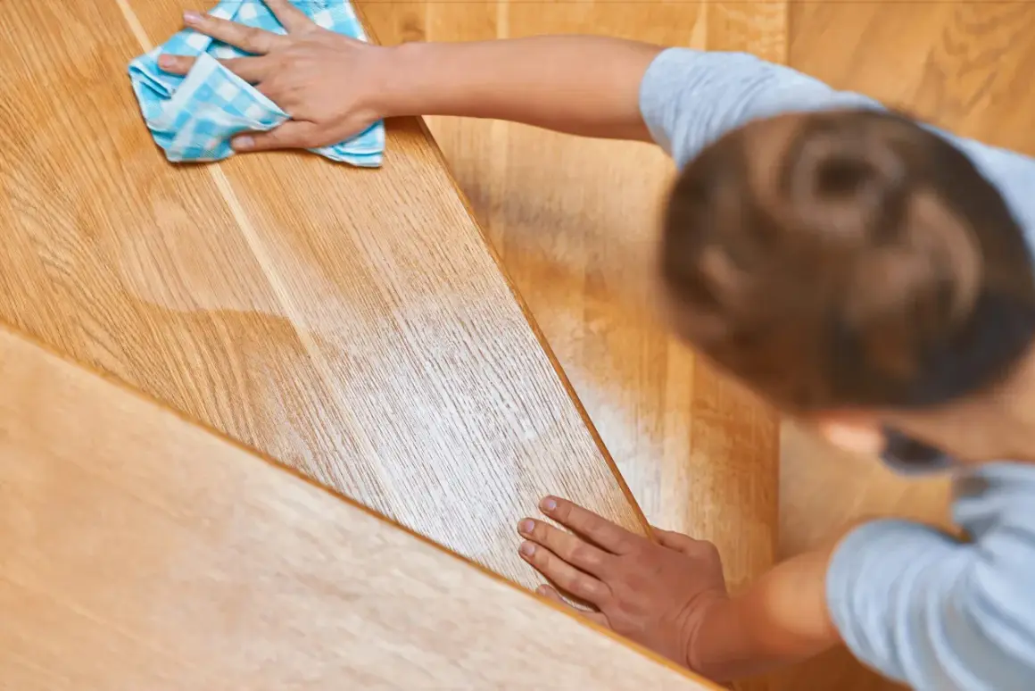 Une main nettoie un escalier en bois verni encrassé avec un chiffon bleu à carreaux.