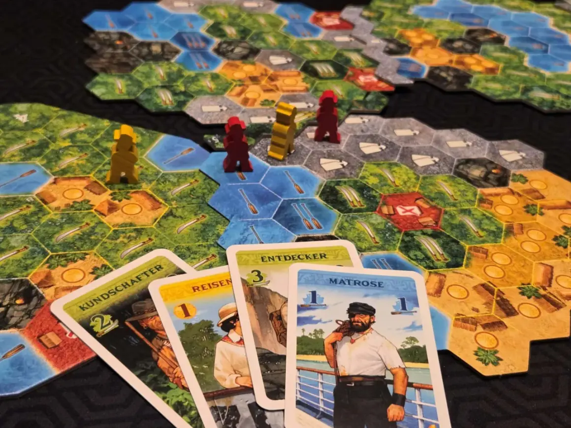 Spielbrett mit Figuren und Karten. Die Karten "KUNDSCHAFTER", "REISEN", "ENTDECKER" und "MATROSE" sind ausgelegt. Eine deutsche Spielepreis Liste könnte diese Elemente zeigen.