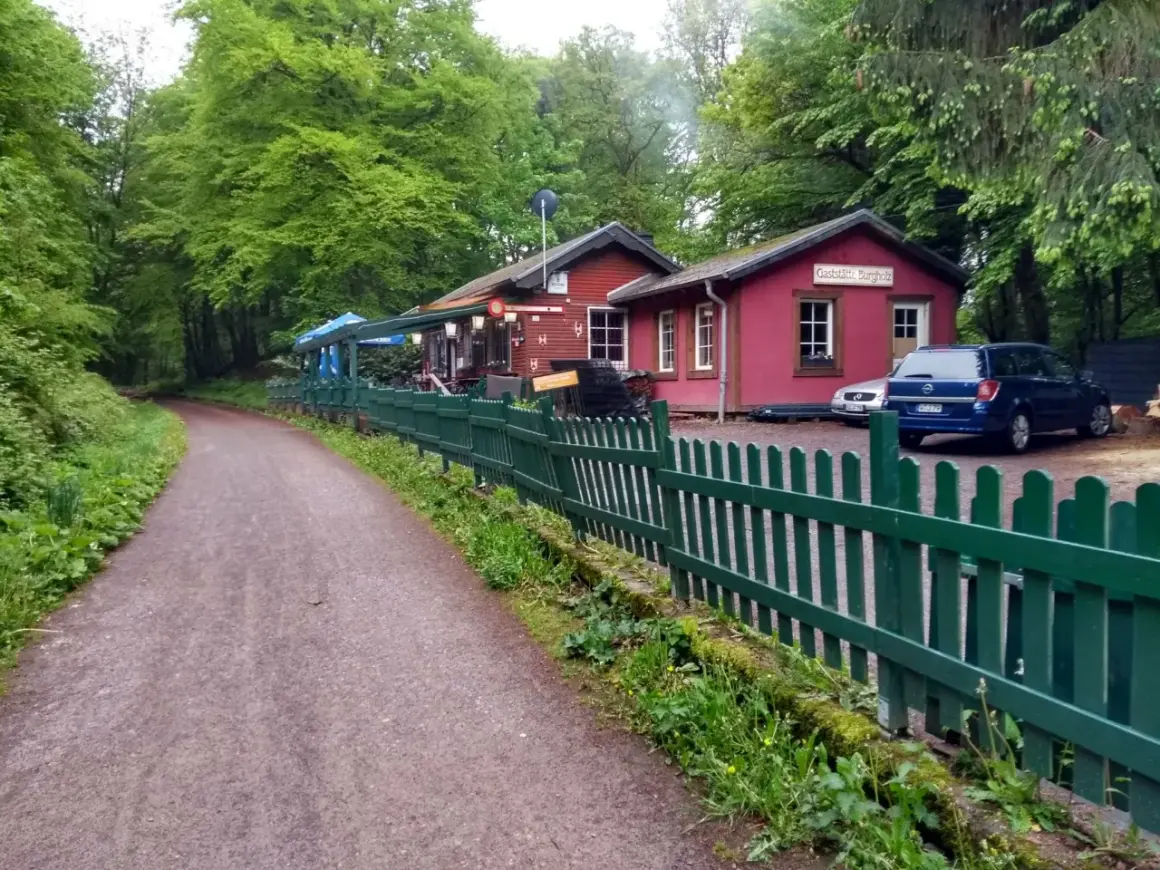 Der Weg zum Bahnhof Burgholz führt vorbei an einer roten Gaststätte mit grünen Zäunen und Autos.