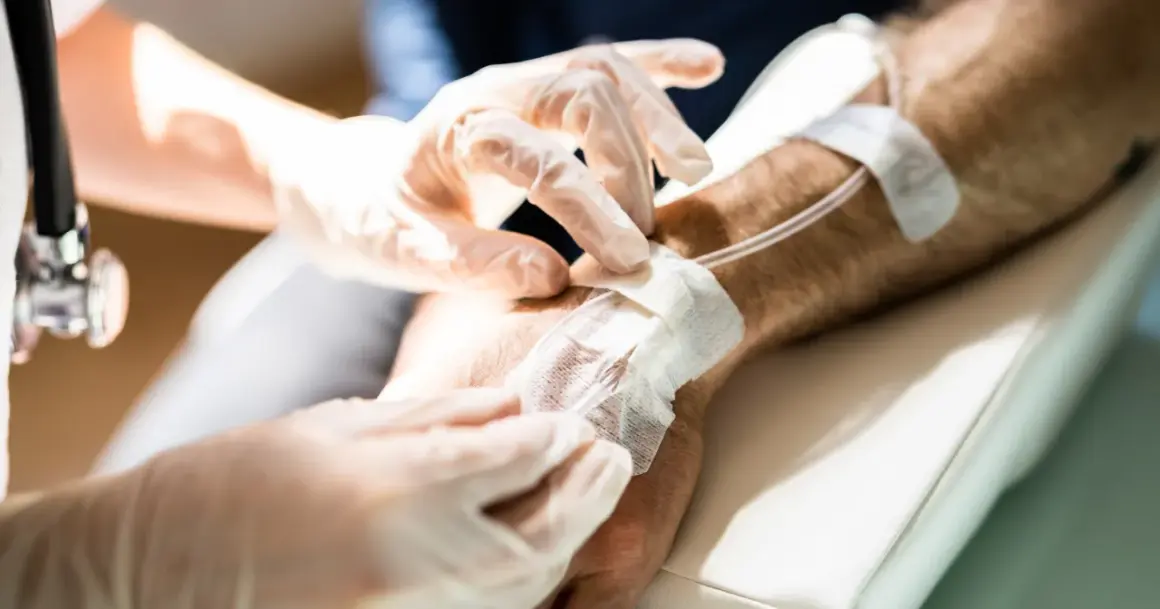 Ręce w rękawiczkach zakładają opatrunek na wenflon. Pacjent przechodzi chemioterapię, która bywa trudna, ale każdy cykl przybliża do zdrowia.
