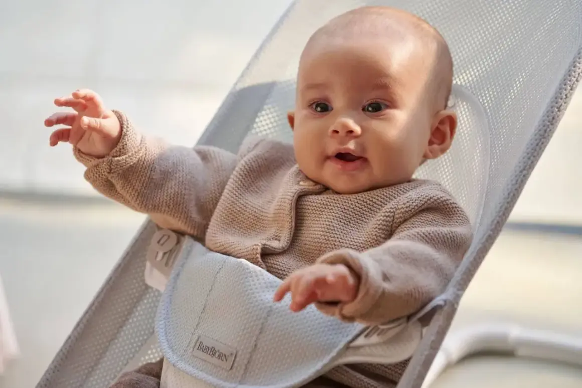 Uśmiechnięte niemowlę w beżowym sweterku siedzi w leżaczku BabyBjörn. Ten leżaczek jest idealny od pierwszych miesięcy życia dziecka.