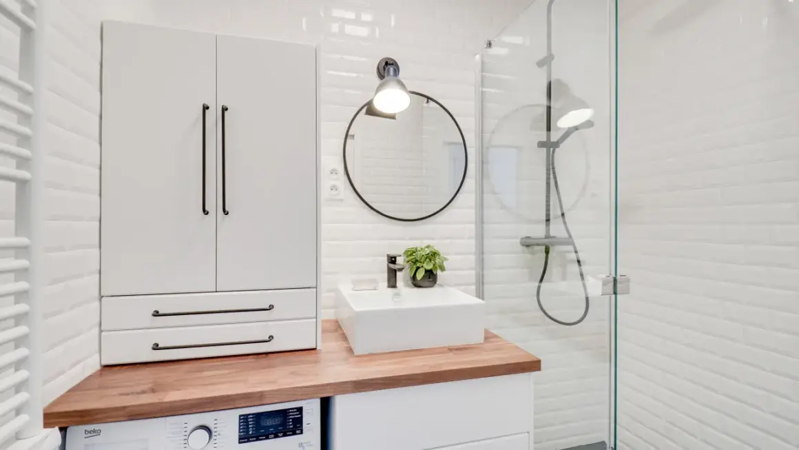 Coin buanderie dans salle de bain moderne avec machine à laver, lavabo, miroir rond et douche vitrée.