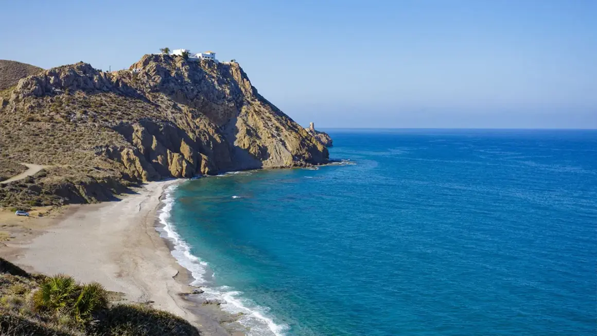Costa rocosa con una playa de arena dorada y aguas turquesas. En lo alto del acantilado, casas blancas y un faro vigilan la costa. Un lugar idílico para disfrutar del sopalmo playa.
