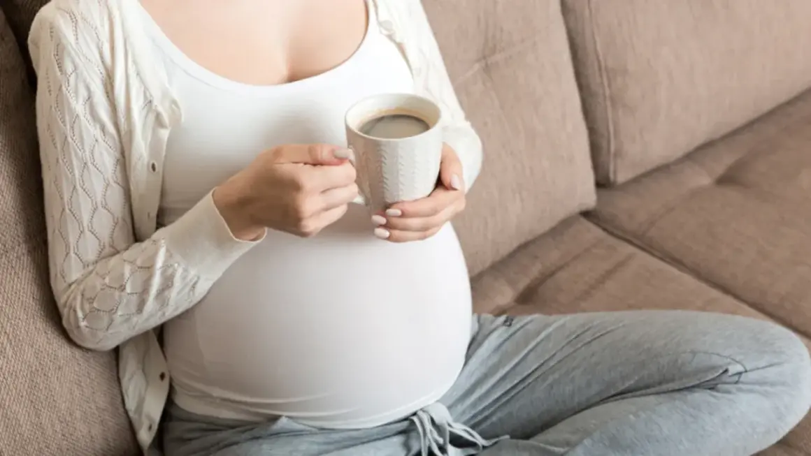 En gravid kvinna sitter på en soffa och håller en mugg med kaffe.
